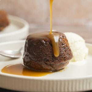 A Sourdough Sticky Date Pudding topped with warm caramel sauce pours over it, served on a plate with a scoop of vanilla ice cream and a spoon beside it.