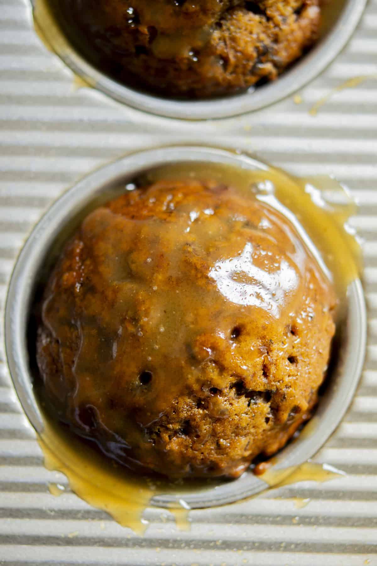 Close-up of Sourdough Sticky Date Puddings in a muffin tin, topped with a shiny layer of caramel sauce. The moist cake glistens as sauce drips onto the ridged metal surface.