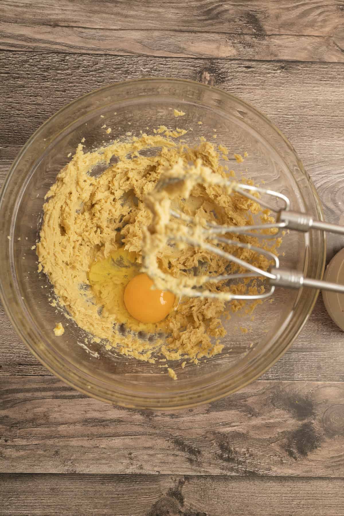 A glass mixing bowl on a wooden surface contains cookie dough with a raw egg on top. An electric hand mixer with batter on the beaters rests inside the bowl.