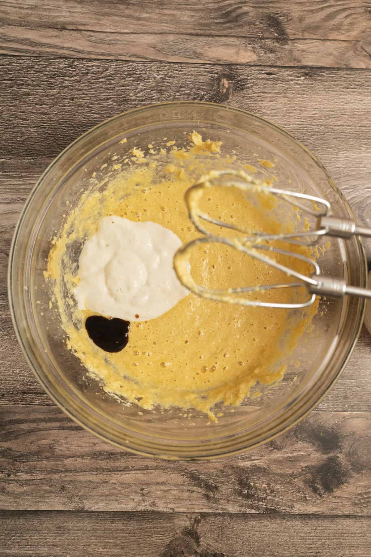 A glass mixing bowl on a wooden surface contains a yellow batter with a dollop of white mixture and a splash of dark liquid. An electric hand mixer with two beaters rests on the bowls edge.