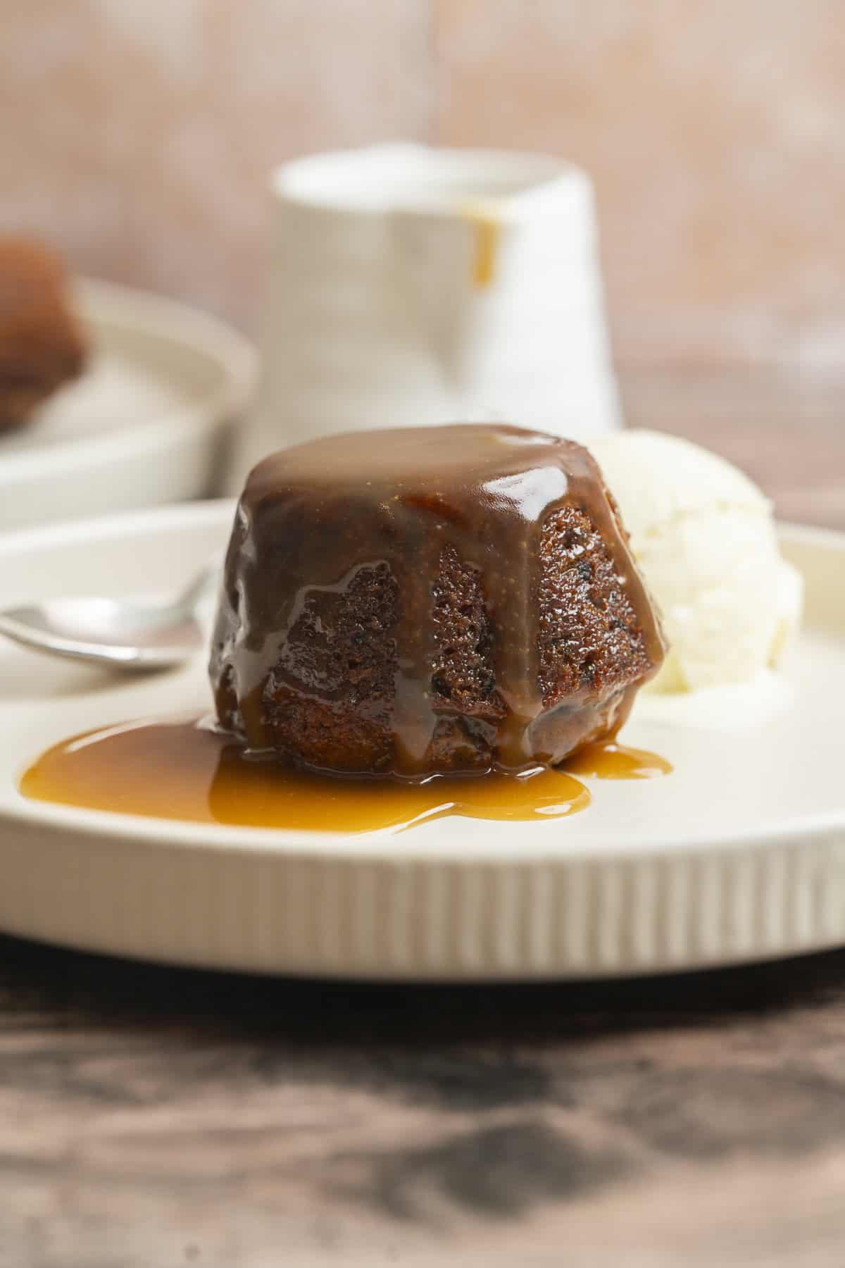 A Sourdough Sticky Date Pudding covered in caramel sauce sits on a plate next to a scoop of vanilla ice cream, with a spoon and a blurred jug in the background.