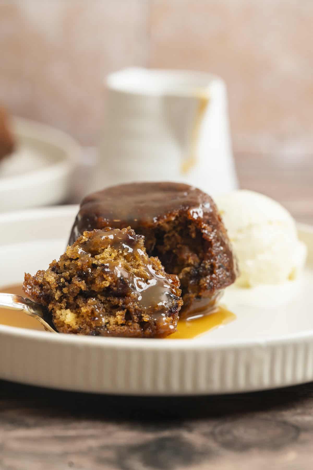 A close-up of Sourdough Sticky Date Puddings with caramel sauce, served with vanilla ice cream on a white plate. A fork holds a bite of the dessert, and a small jug is blurred in the background.