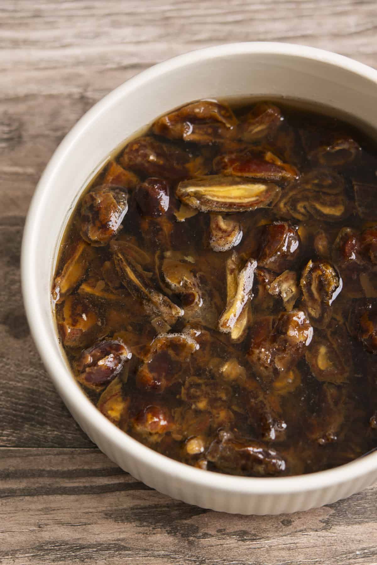 A white bowl filled with chopped dates soaking in water, placed on a wooden surface. The dates appear soft and dark brown, with some pieces floating in the liquid.