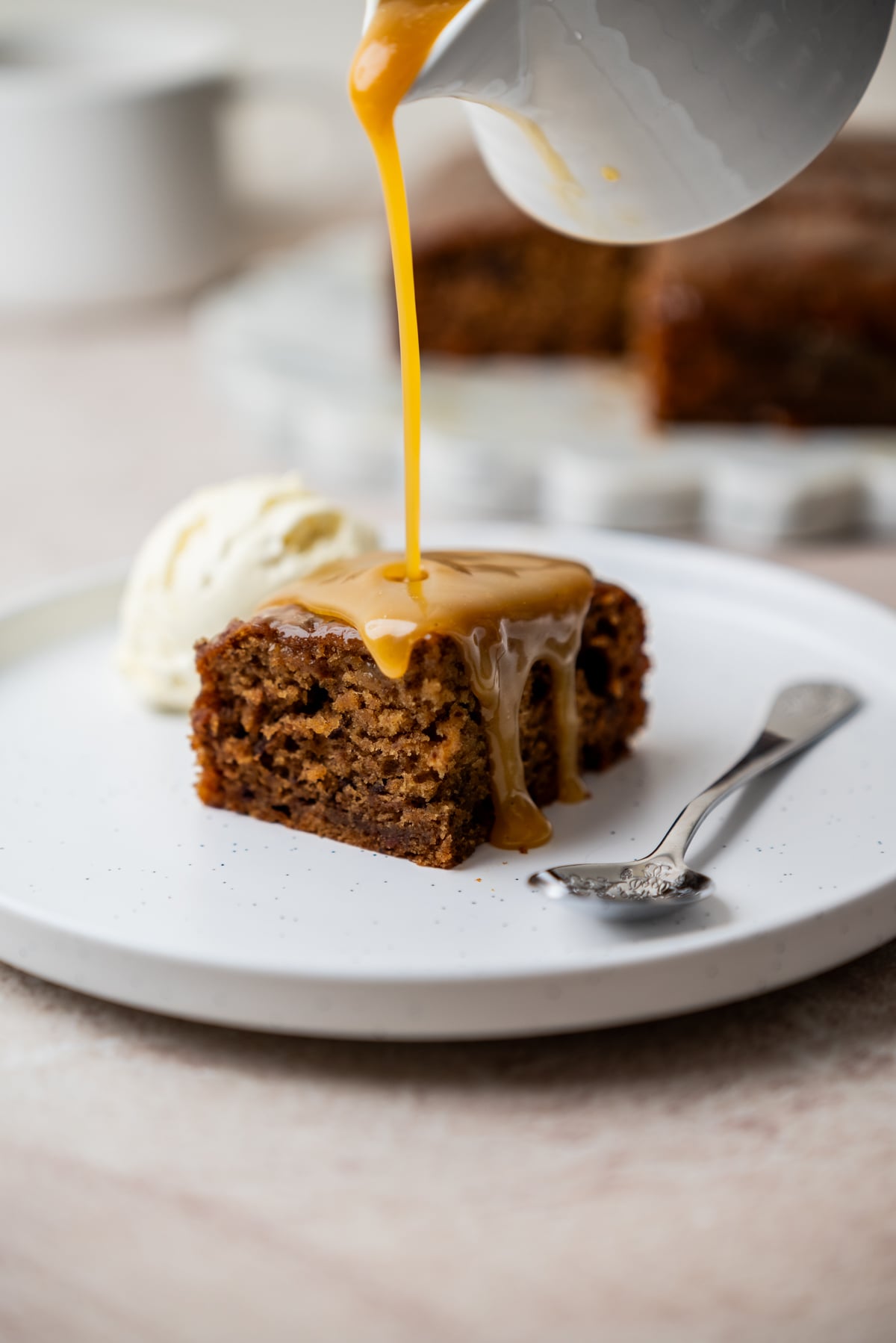 A slice of Sourdough Sticky Date Pudding with caramel sauce being poured over it, served with a scoop of vanilla ice cream on a white plate, next to a spoon.