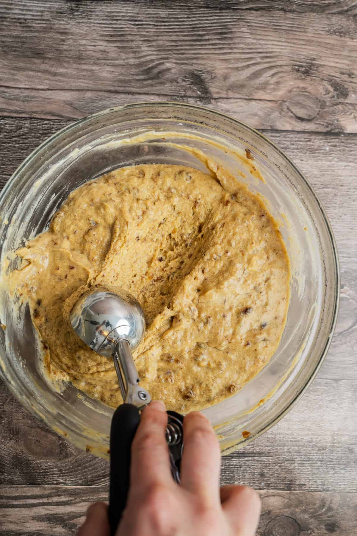 A hand holds an ice cream scoop over a glass bowl filled with cookie dough mixture, set on a wooden surface.
