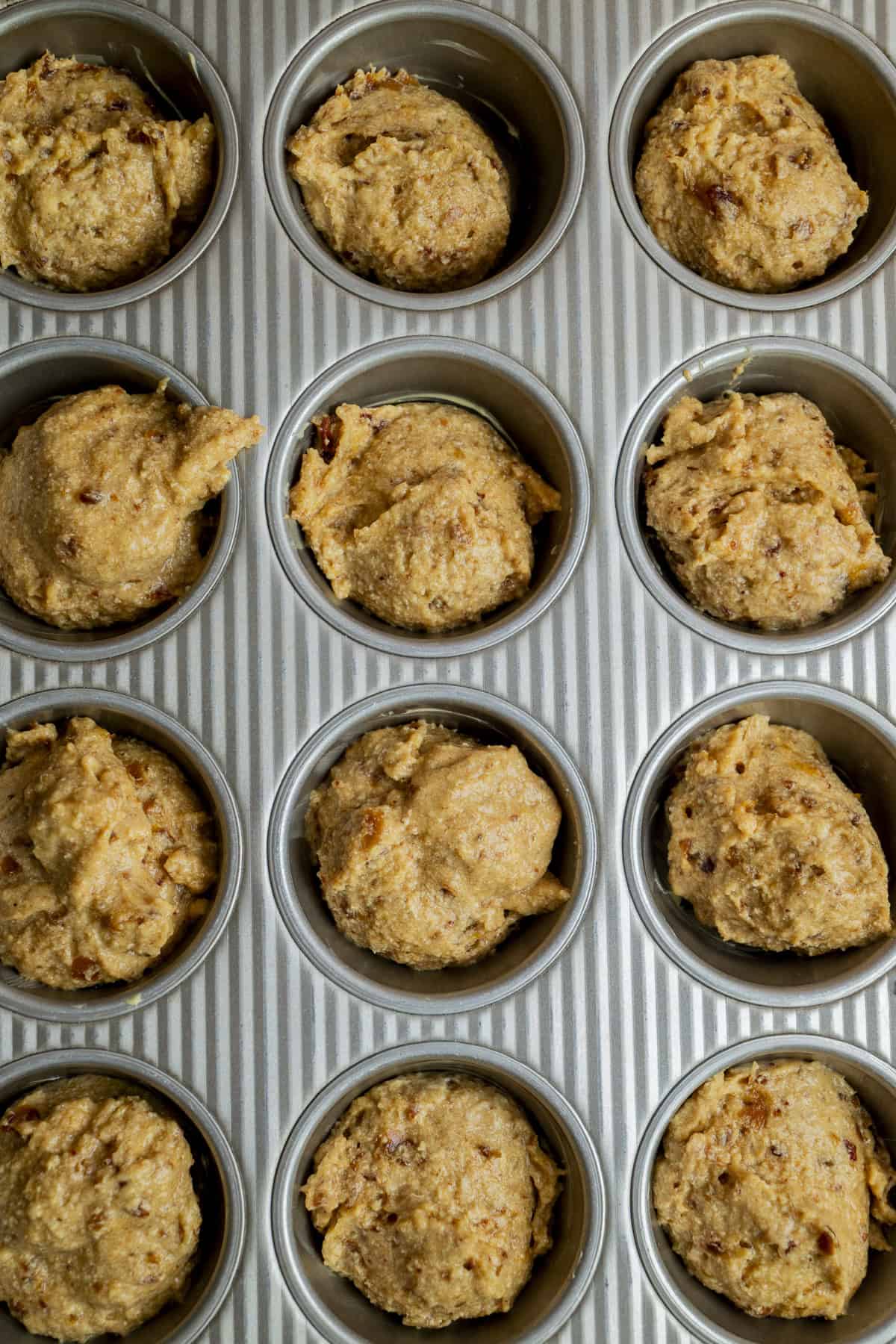 A muffin tin filled with raw muffin batter, divided into twelve sections, ready to be baked. The batter is light brown with visible bits mixed throughout.