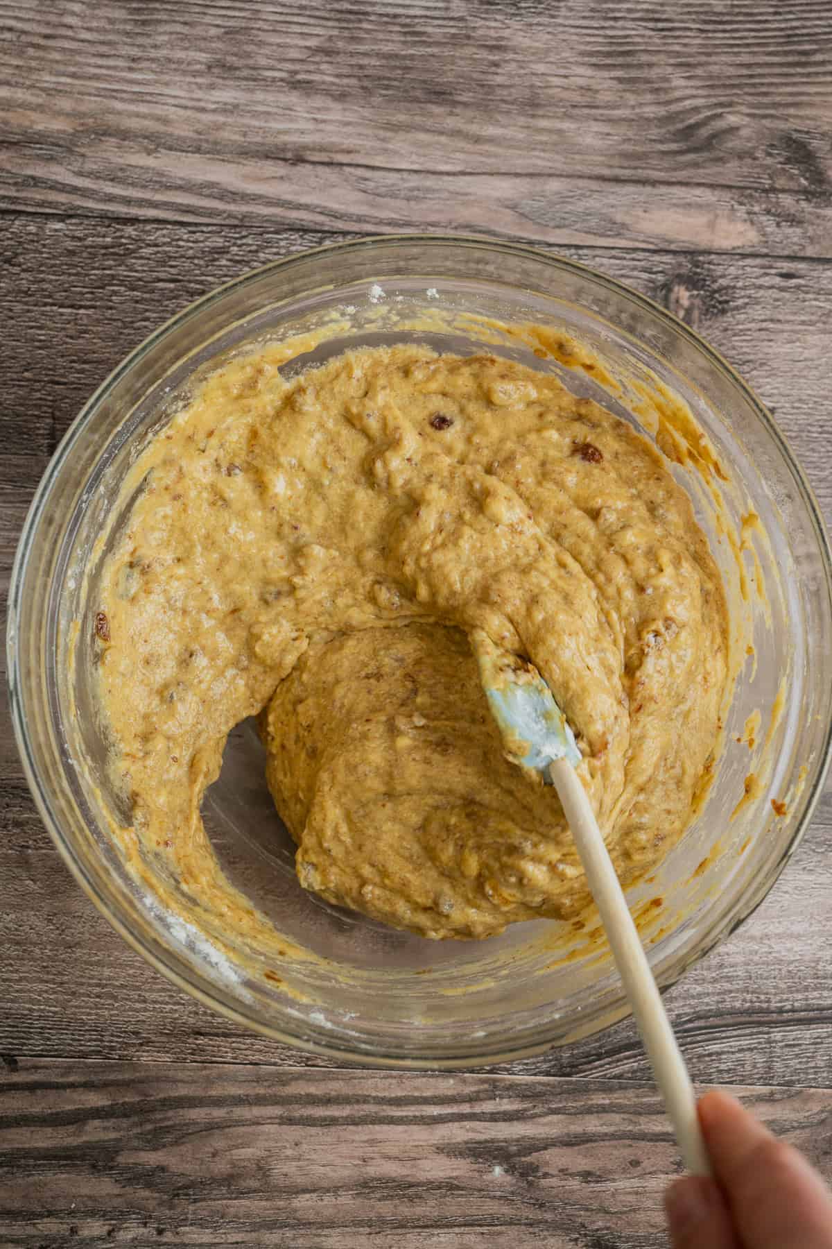 A hand holding a spatula mixes thick, yellowish batter with visible raisins in a glass bowl on a wooden surface.