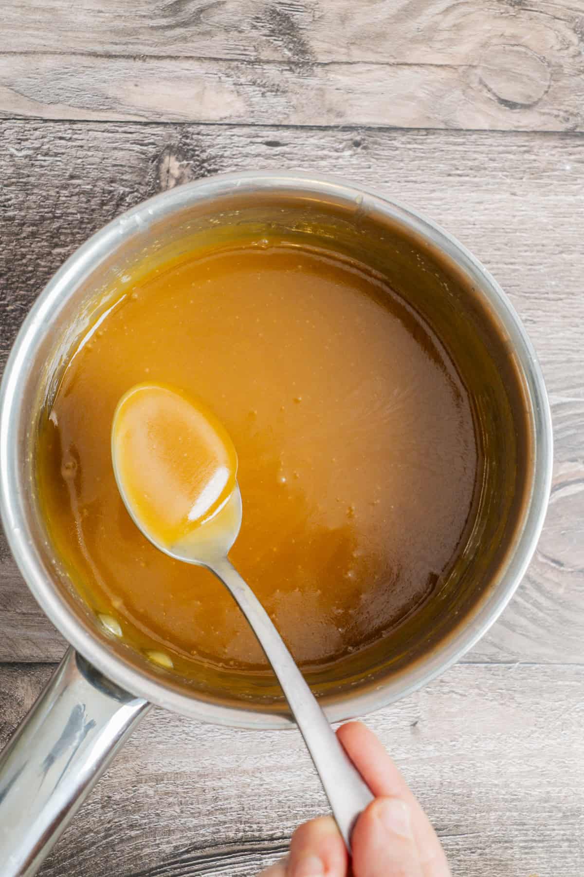 A hand holds a spoon scooping golden caramel sauce from a saucepan, set on a wooden surface. The caramel is smooth and glossy, filling most of the pan.
