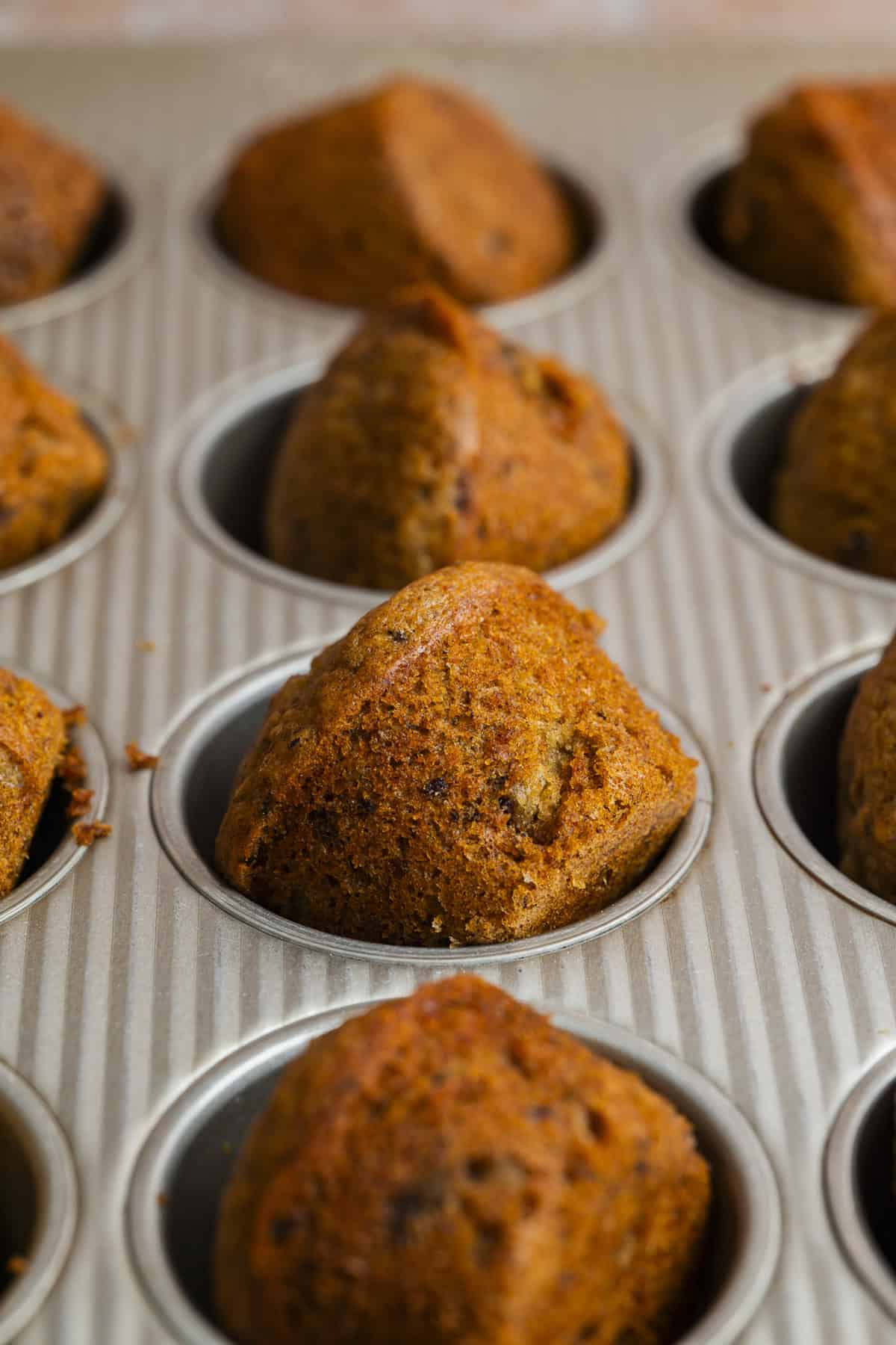 A close-up of freshly baked date puddings in a metal muffin tin, with golden-brown tops and a slightly uneven texture, suggesting they contain nuts or fruit.
