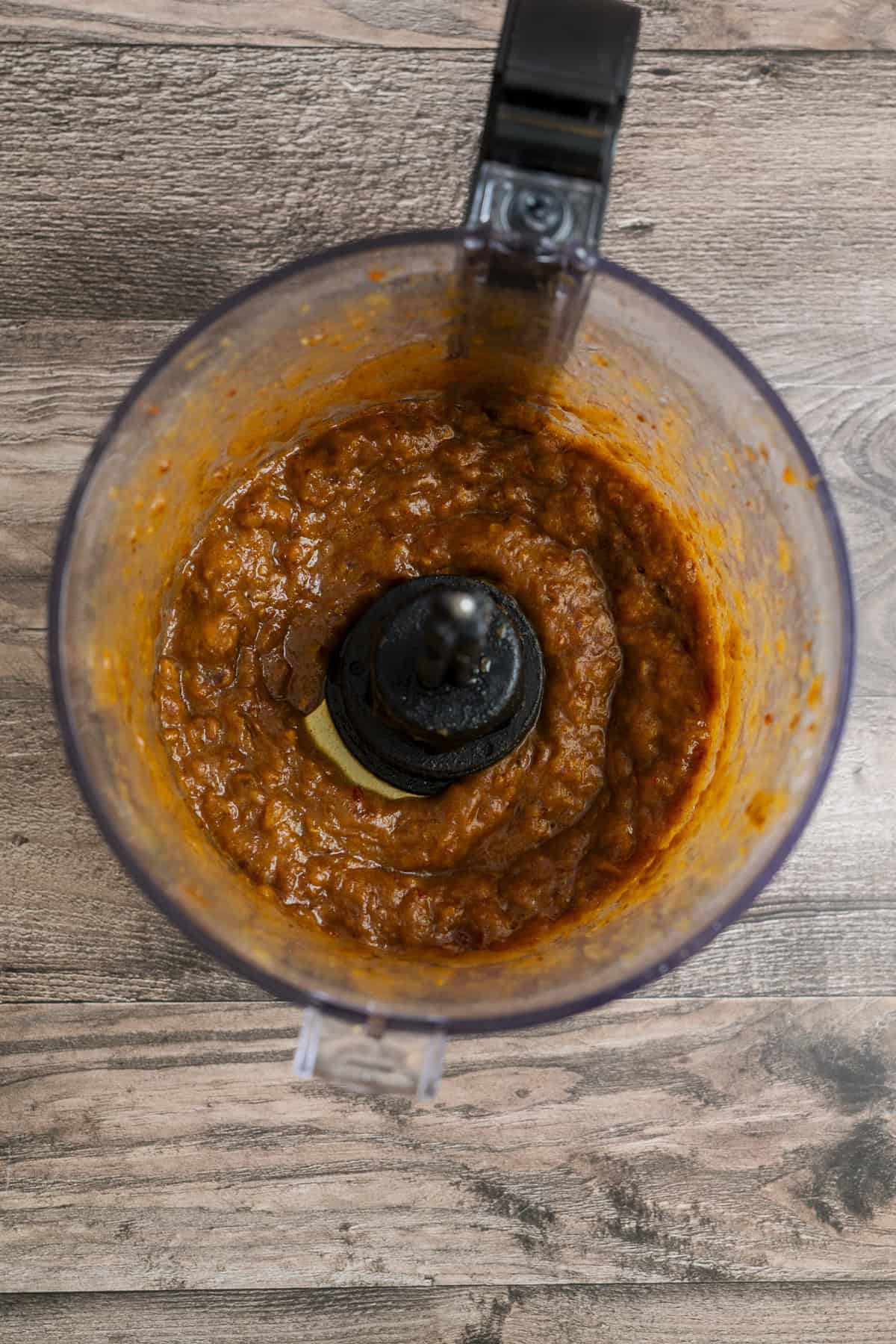 A top-down view of a food processor containing a thick, orange-brown sauce or paste, set on a wooden surface. The sauce appears to be freshly blended.