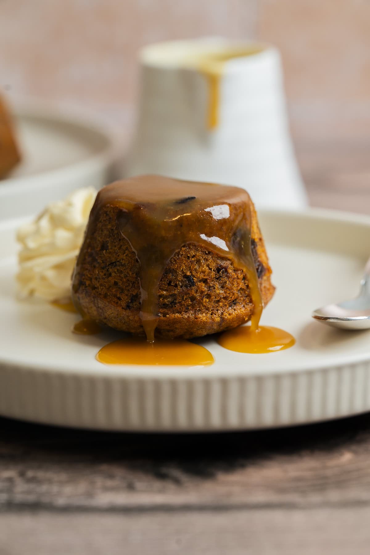 A sticky toffee pudding topped with caramel sauce sits on a white plate, next to a swirl of whipped cream. A spoon and a small white jug of sauce are in the background.