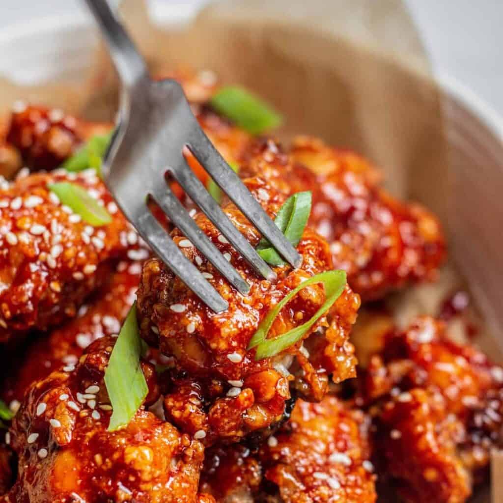 A fork holds a piece of crispy, saucy fried chicken garnished with sesame seeds and sliced green onions, with more pieces in the background.