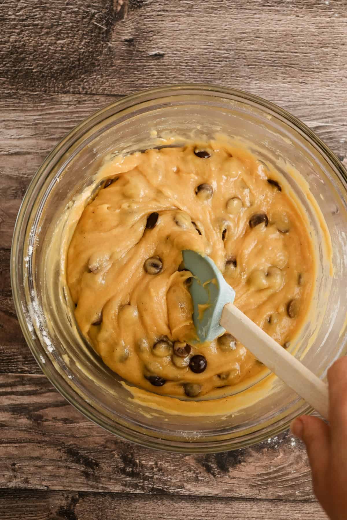 A hand stirs chocolate chip cookie dough in a clear glass bowl with a blue spatula on a wooden surface.