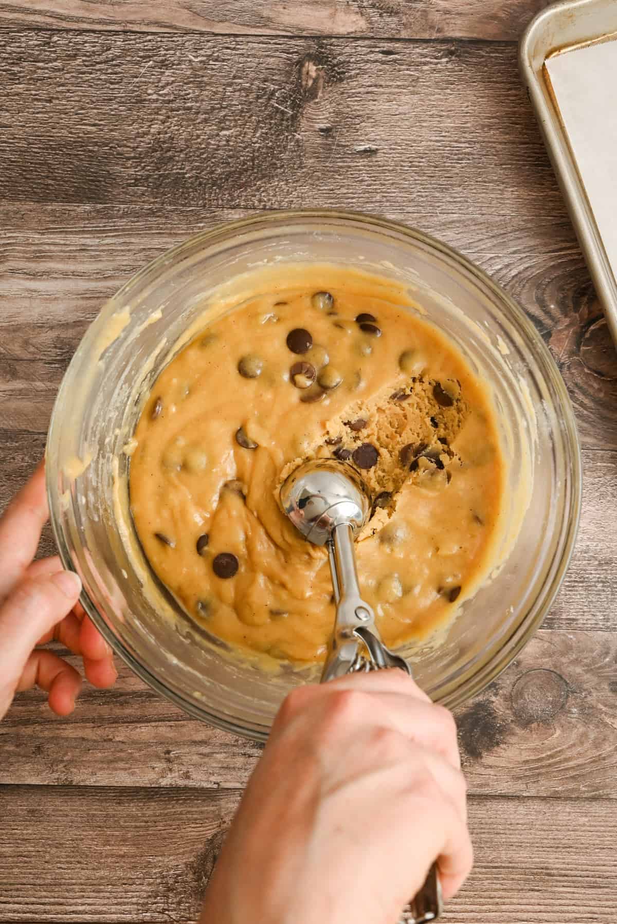 A hand holds a cookie scoop over a glass bowl of chocolate chip cookie dough on a wooden surface, with a baking sheet lined with parchment paper nearby.