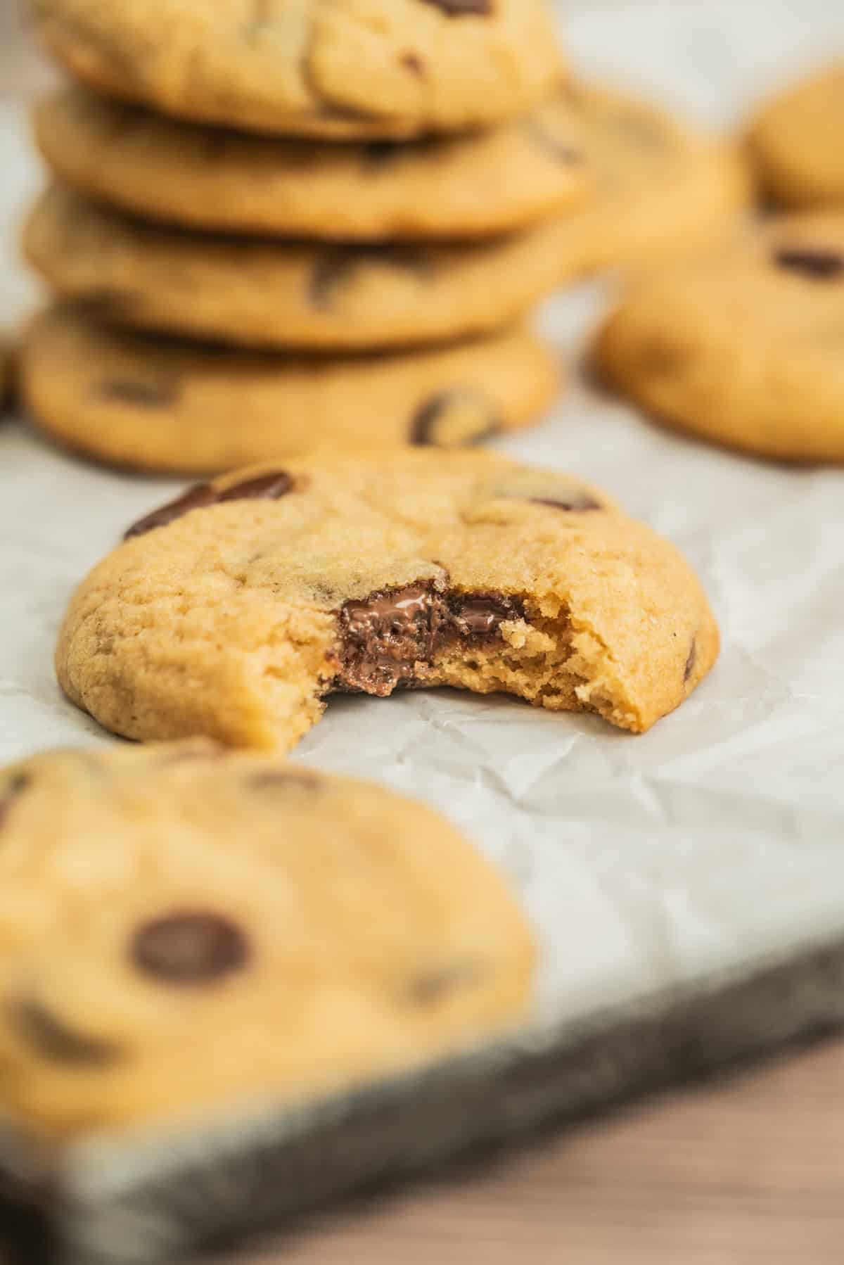 A close-up of a sourdough chocolate chip cookie with a bite taken out, revealing a gooey chocolate center. Other cookies are stacked and scattered in the background on parchment paper.