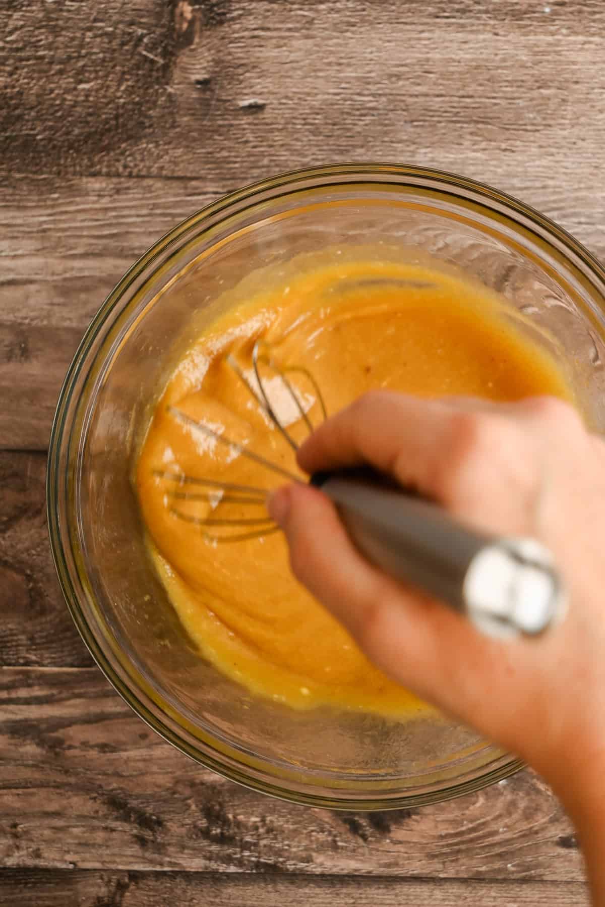 A hand uses a metal whisk to mix a thick, yellow-orange batter in a clear glass bowl on a wooden surface.