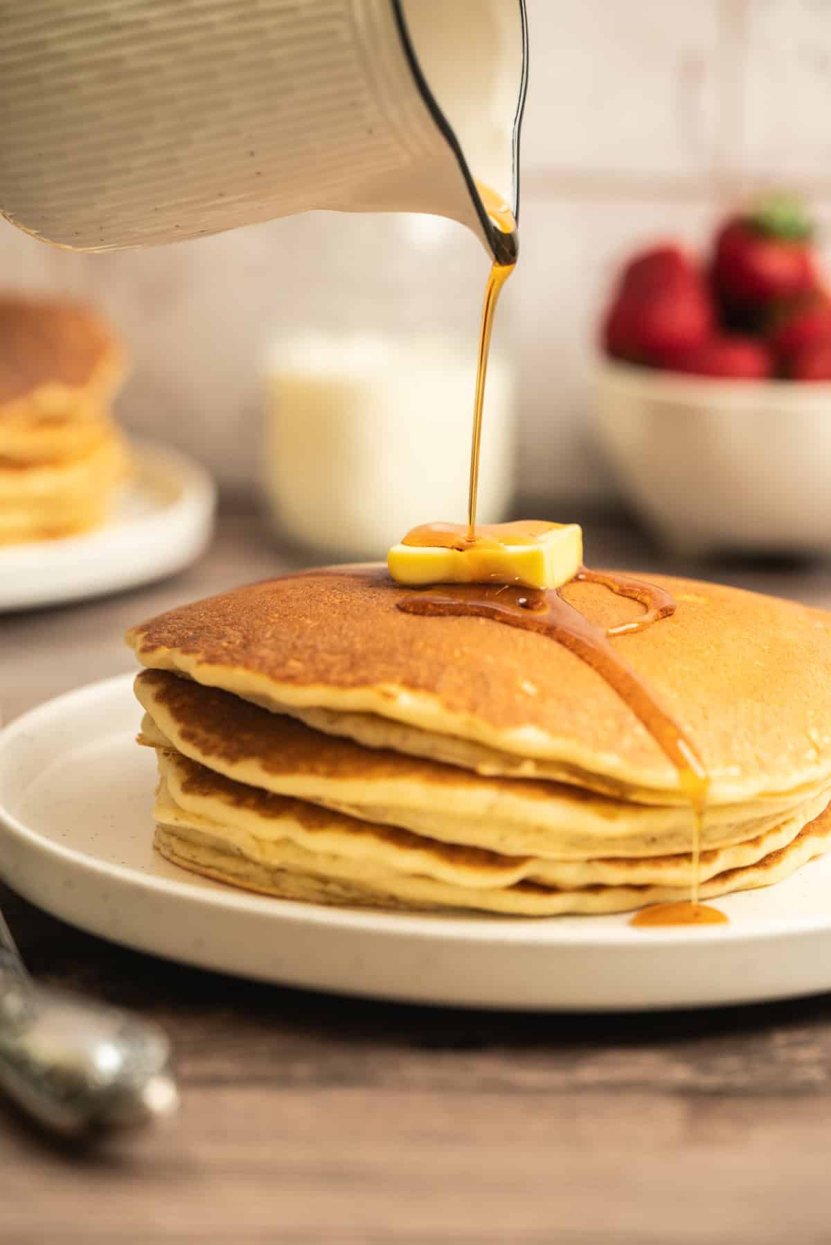 A stack of sourdough pancakes topped with a pat of butter is being drizzled with syrup from a pitcher. In the background, there are strawberries in a bowl and another stack of pancakes.