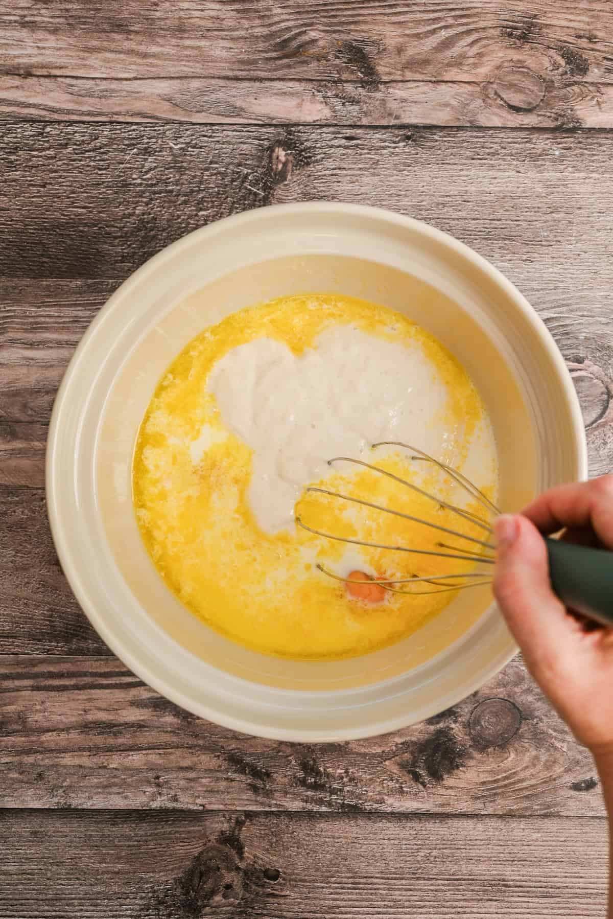 A hand uses a whisk to mix eggs, melted butter, and flour in a beige bowl on a wooden surface.
