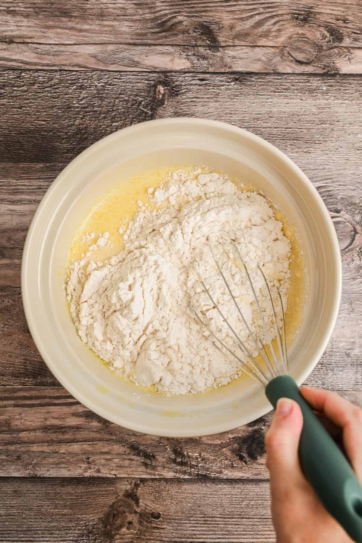 A hand holding a whisk mixes flour and wet ingredients in a large beige mixing bowl on a wooden surface.
