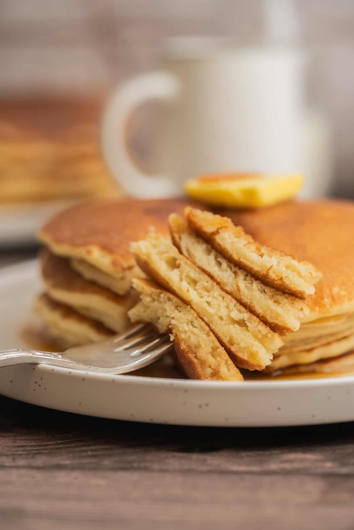 A close-up of a stack of sourdough starter pancakes topped with a pat of butter. A fork holds a bite-sized piece of pancakes, showing their fluffy texture. A white mug is blurred in the background.