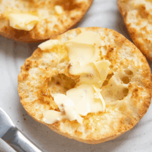 A toasted air fryer English muffin split in half, topped with melting butter, sits on a plate next to a butter knife.