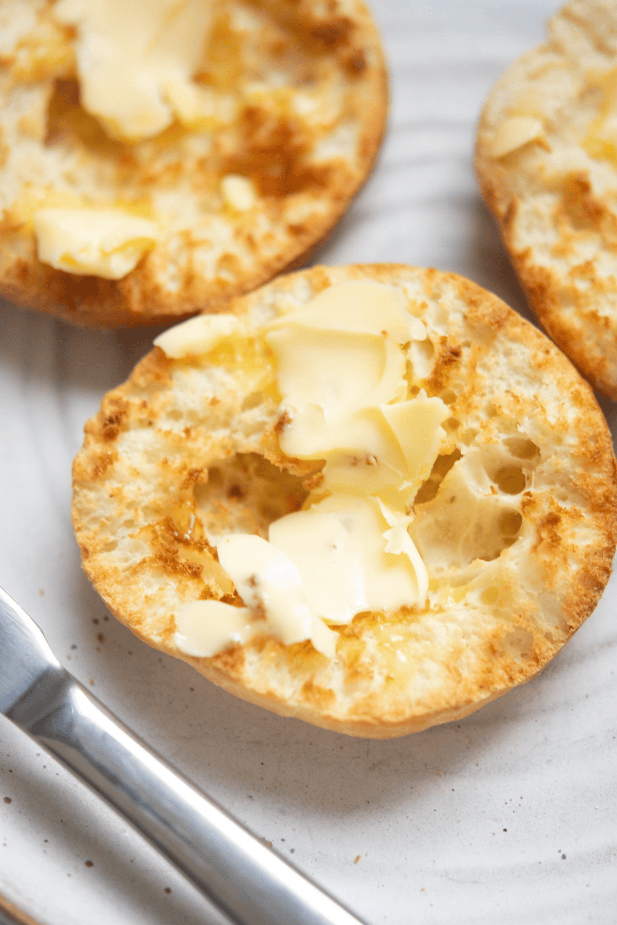 A toasted English muffin split in half, topped with melting butter, sits on a plate next to a butter knife.