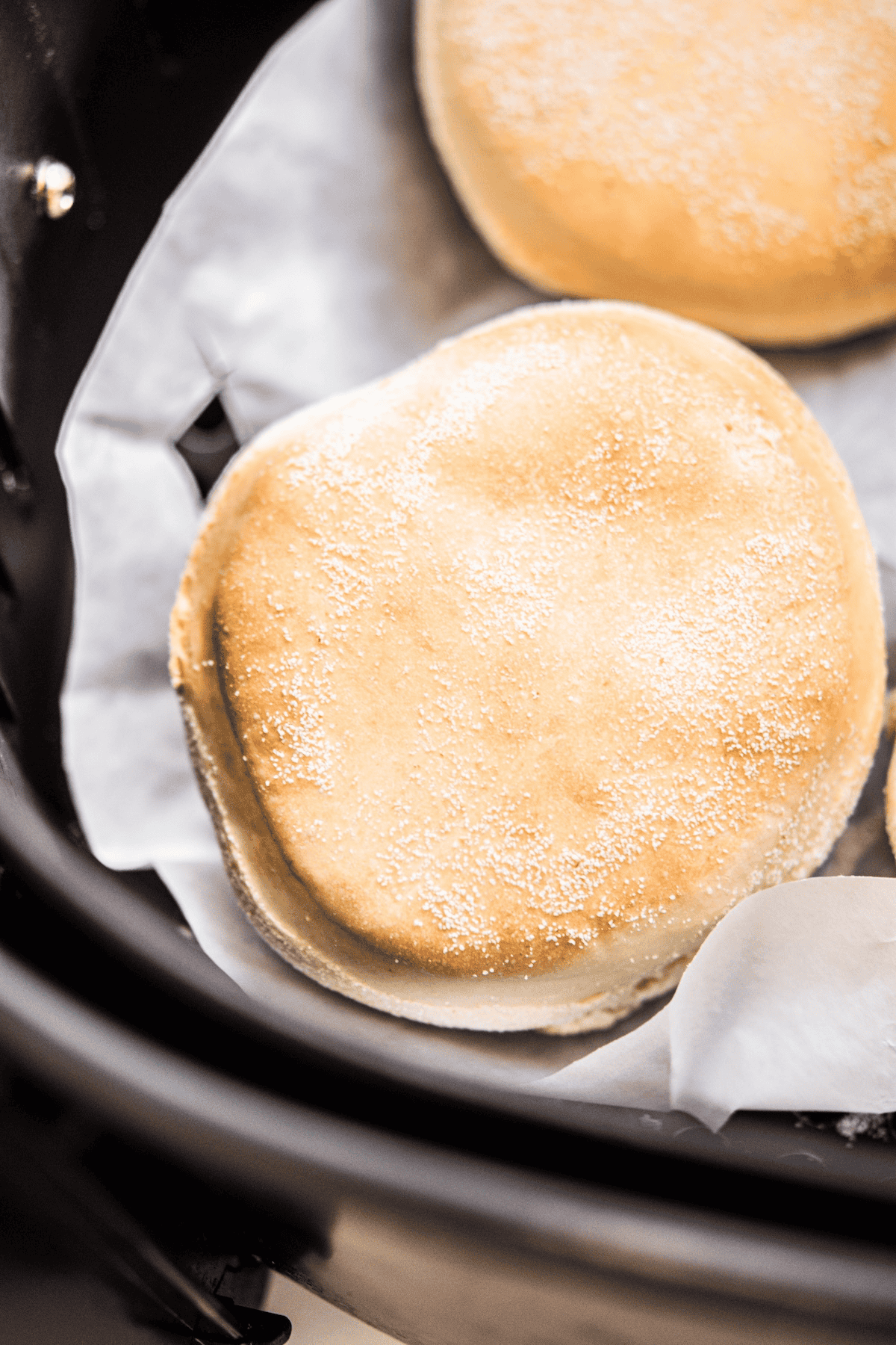 A close-up of a golden brown English muffin on parchment paper inside an air fryer basket.