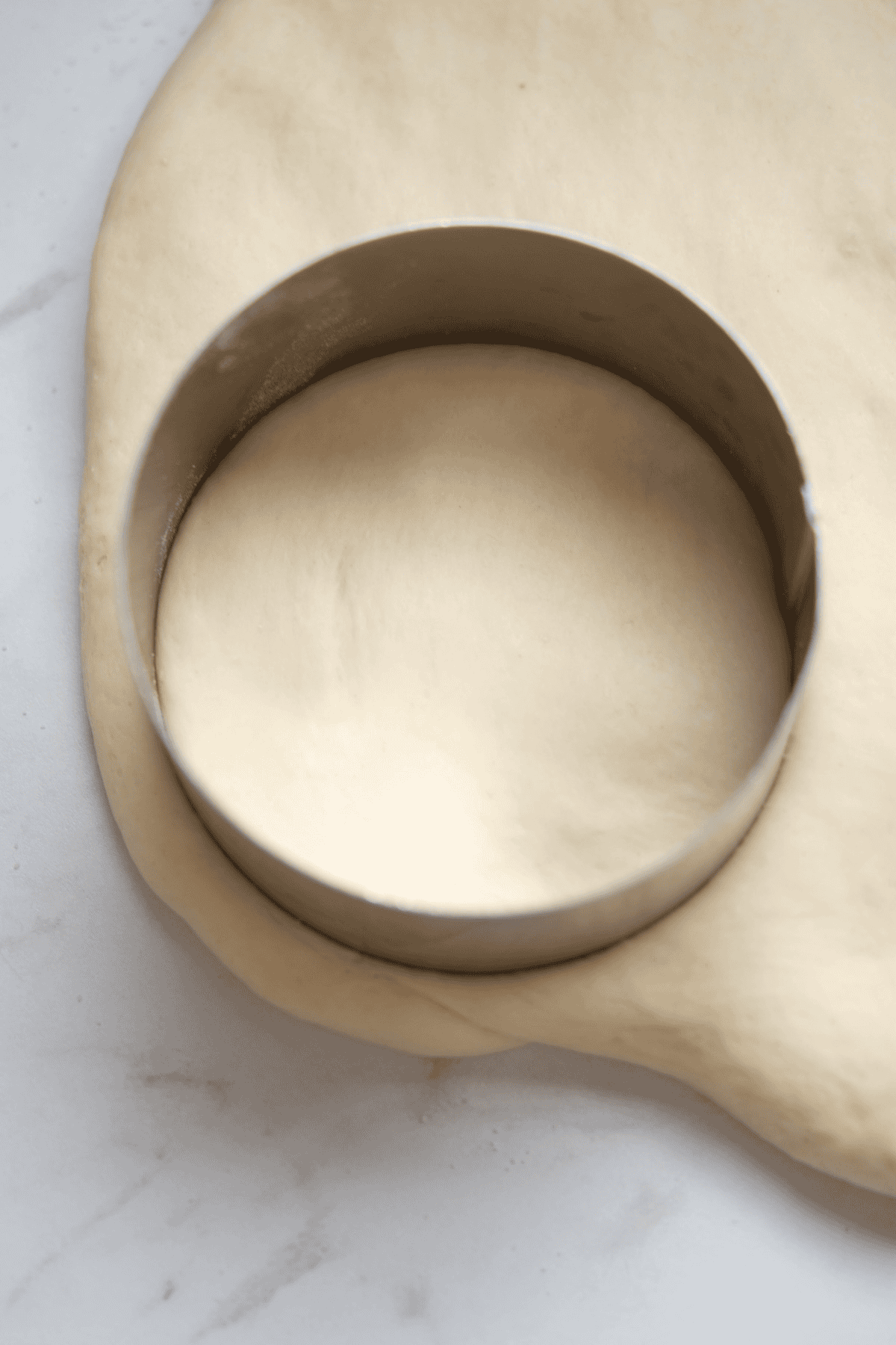 Round metal cutter pressed into rolled-out dough on a marble surface, preparing to cut out a circular shape for baking.