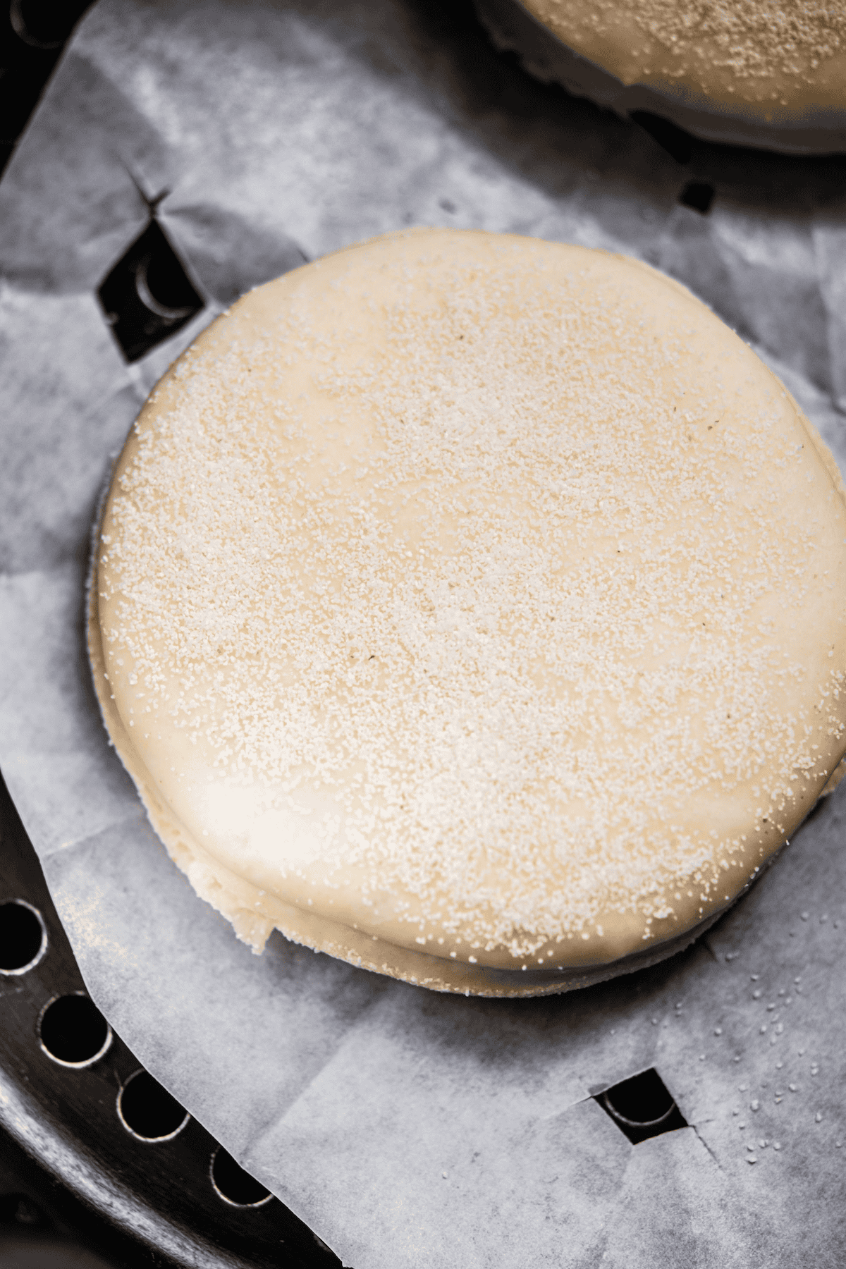 A close-up of an unbaked, round dough disc sprinkled with flour or cornmeal, resting on parchment paper. The surface is smooth, and the parchment paper lines a baking tray beneath the dough.