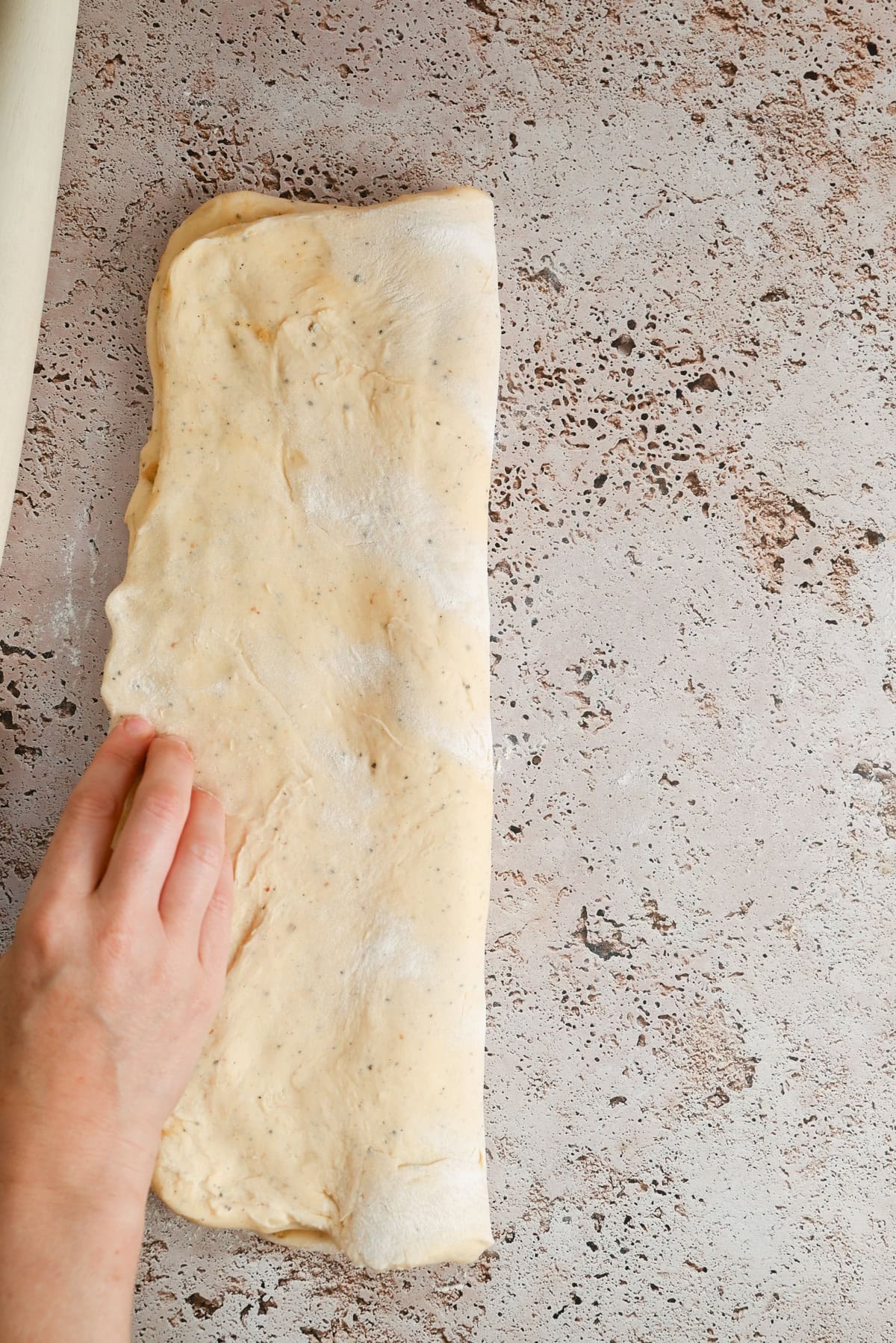 A hand folding a sheet of dough on a speckled countertop, with a rolling pin partially visible in the top left corner.