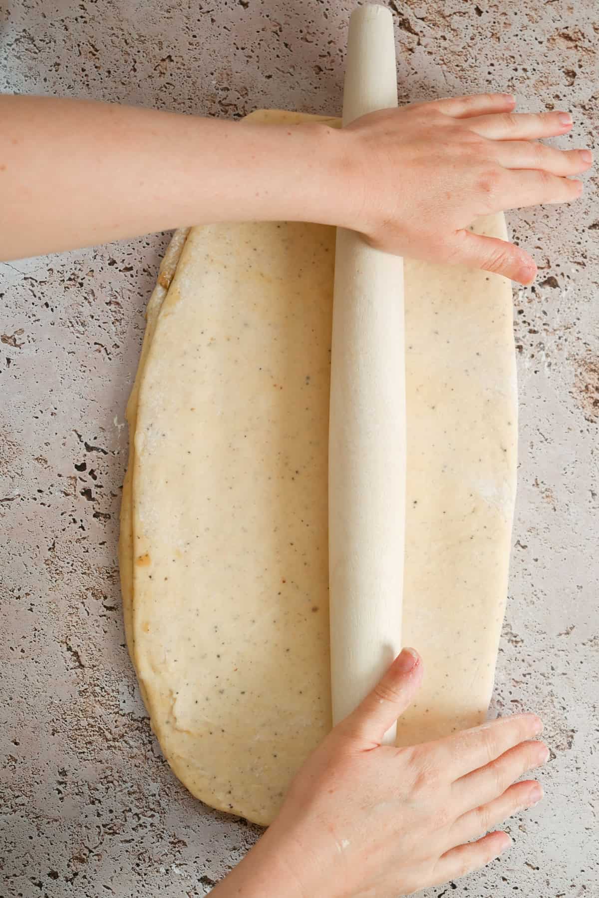 Hands rolling out a rectangle of dough with a rolling pin on a speckled countertop.