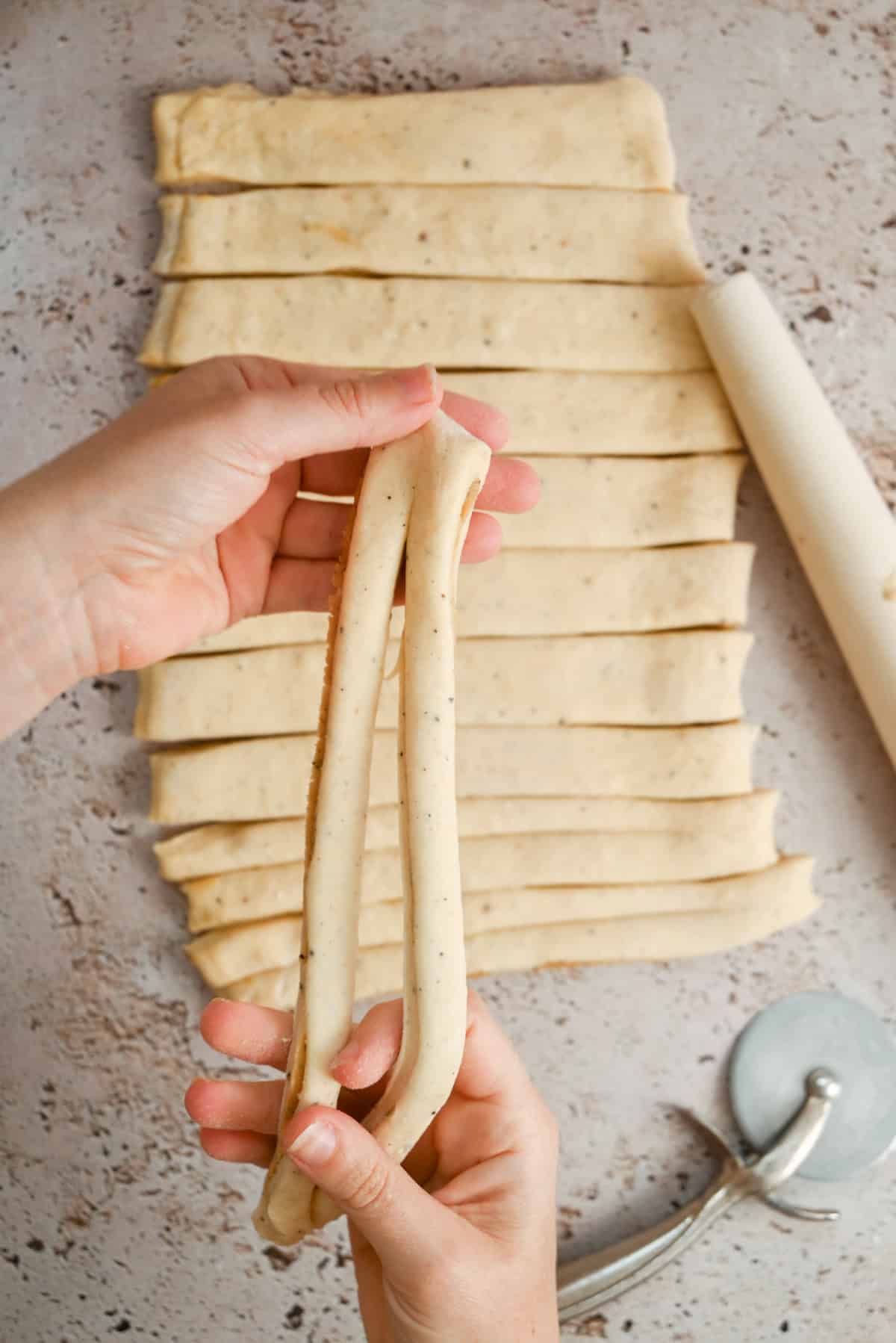 Hands holding two strips of dough, with more strips laid out on a textured surface. A rolling pin and a pastry cutter are nearby. The dough appears freshly cut and ready for further preparation.