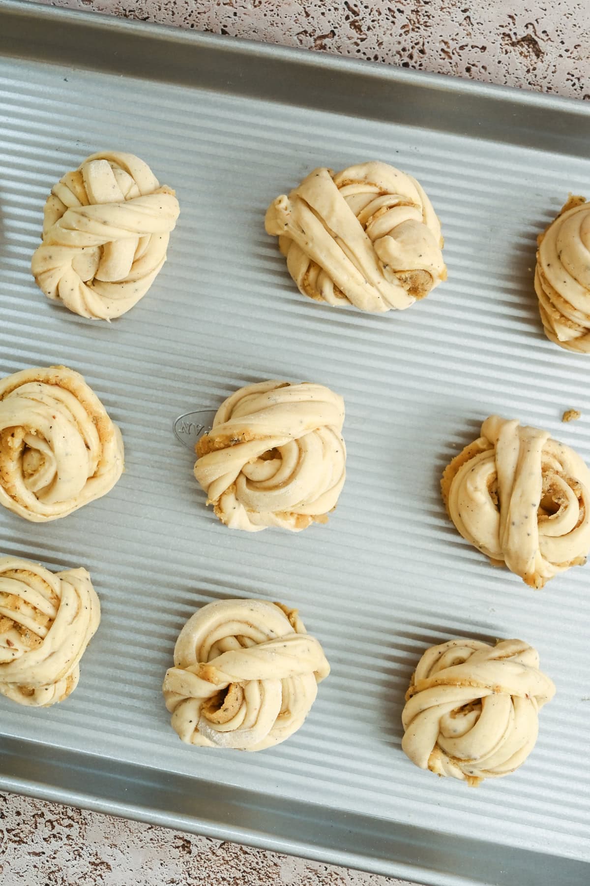 Nine unbaked twisted pastry buns with visible layers and specks of filling are arranged in rows on a silver, ridged baking sheet. The surface beneath the tray is a light, speckled stone.