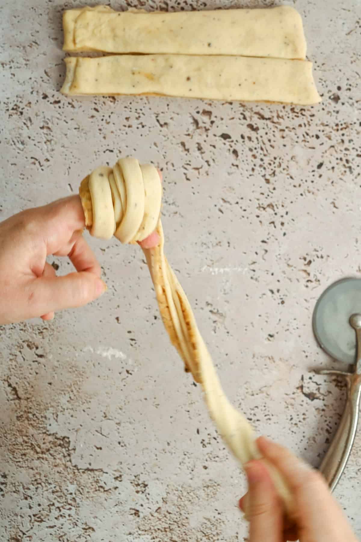 A hand twists a long strip of dough into a knot on a light speckled surface; rectangular strips of dough and a pizza cutter are visible nearby.