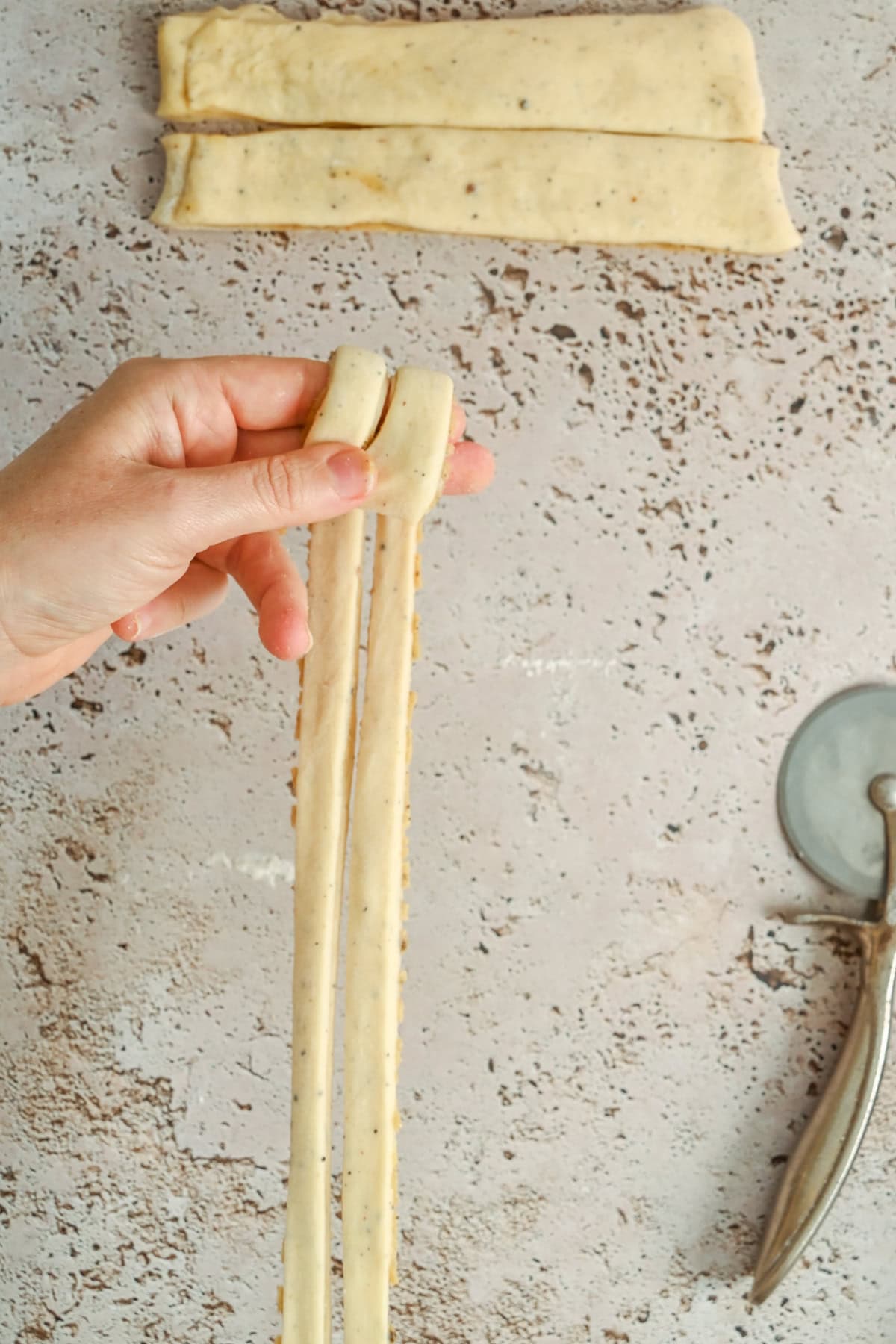 A hand holds two long strips of dough being twisted together, with more dough strips and a pizza cutter on a speckled counter.