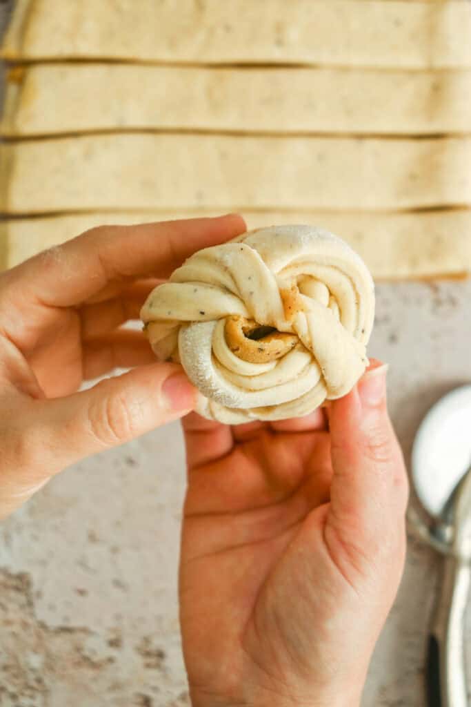 Two hands hold a twisted, unbaked pastry knot in the foreground. In the background, rectangular strips of dough are laid out on a lightly floured surface.