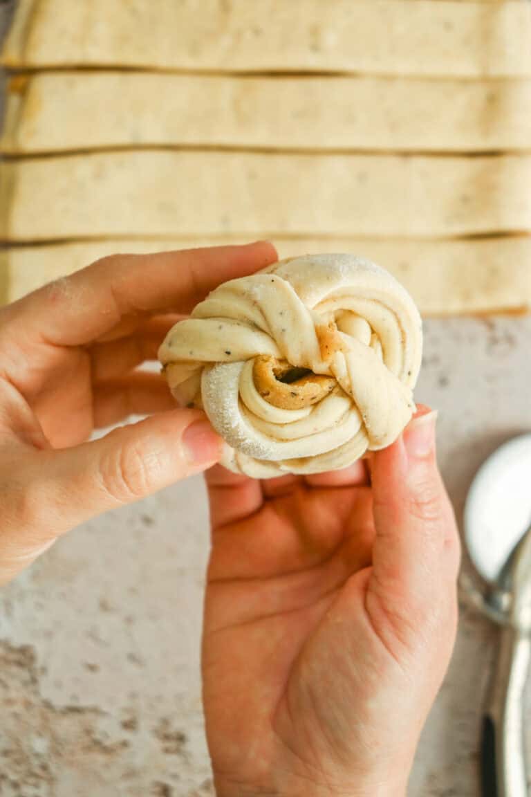 Two hands hold a twisted, unbaked pastry knot in the foreground. In the background, rectangular strips of dough are laid out on a lightly floured surface.