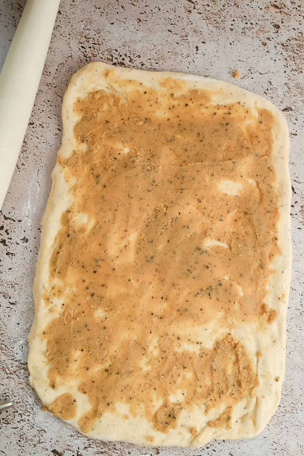 A rectangular slab of dough is spread with a brown, speckled mixture, lying on a textured surface. A rolling pin is partially visible on the left side of the image.