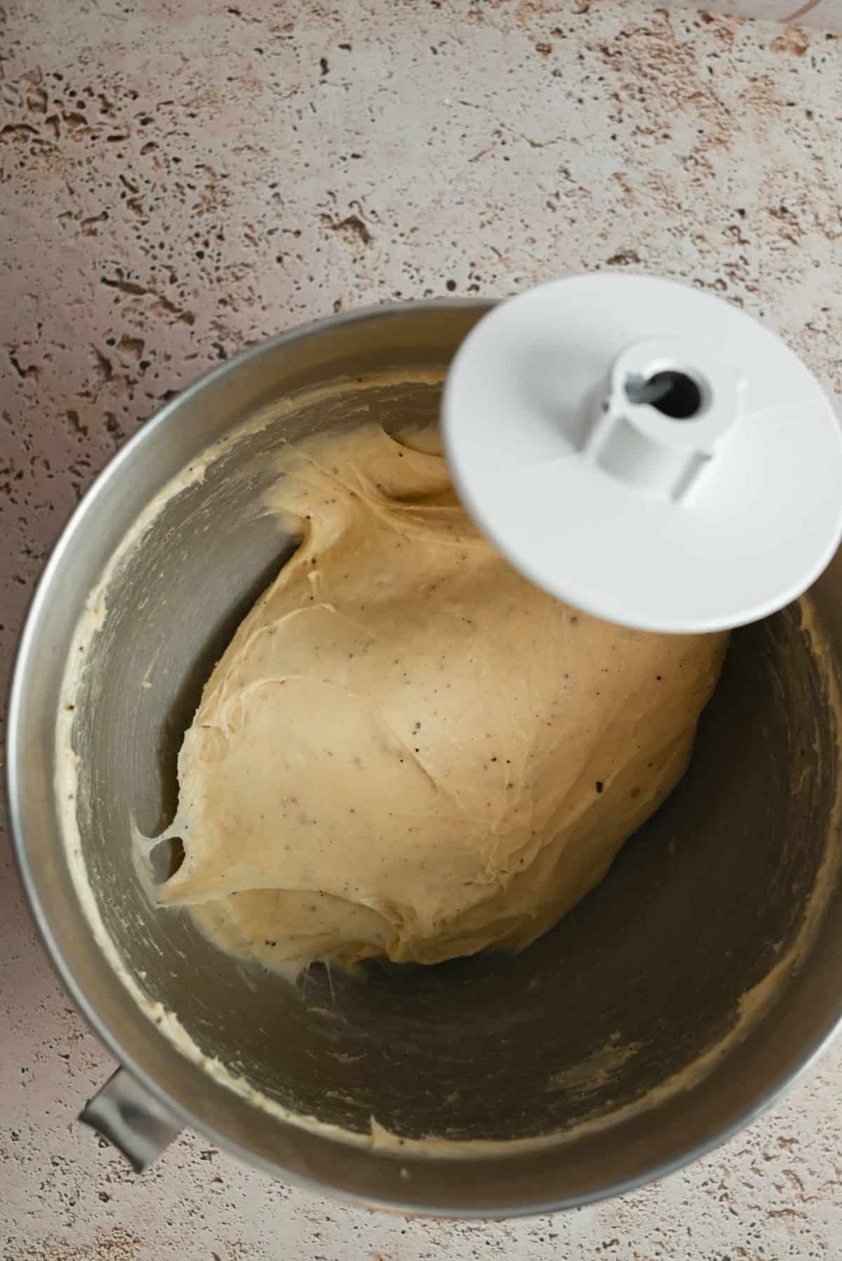 A metal stand mixer bowl with a hook attachment contains smooth, slightly sticky dough, resting on a textured light surface.