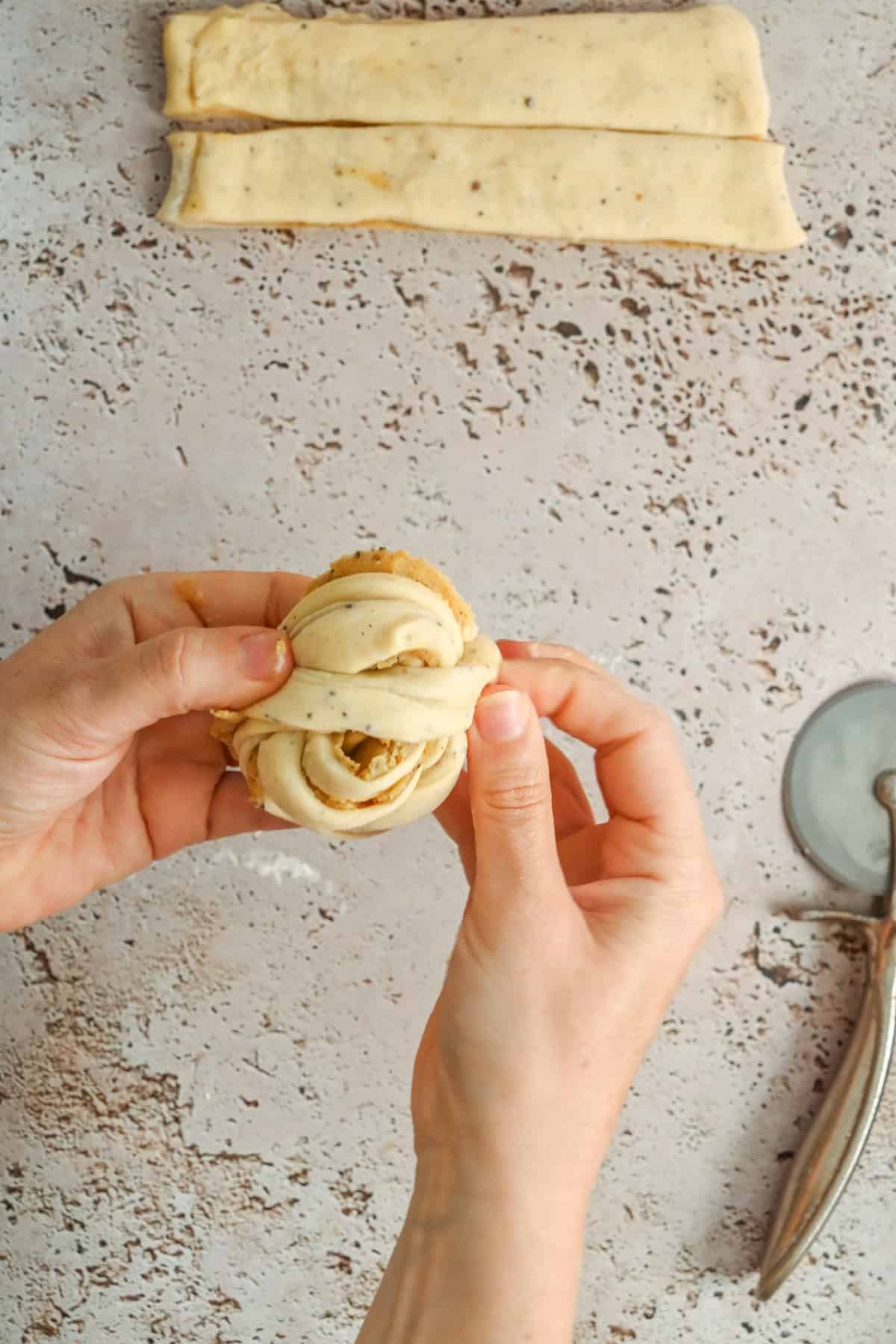 Hands are shaping a strip of dough, likely for a pastry, on a speckled countertop. A pizza cutter and scissors are nearby, with more dough strips visible at the top of the image.