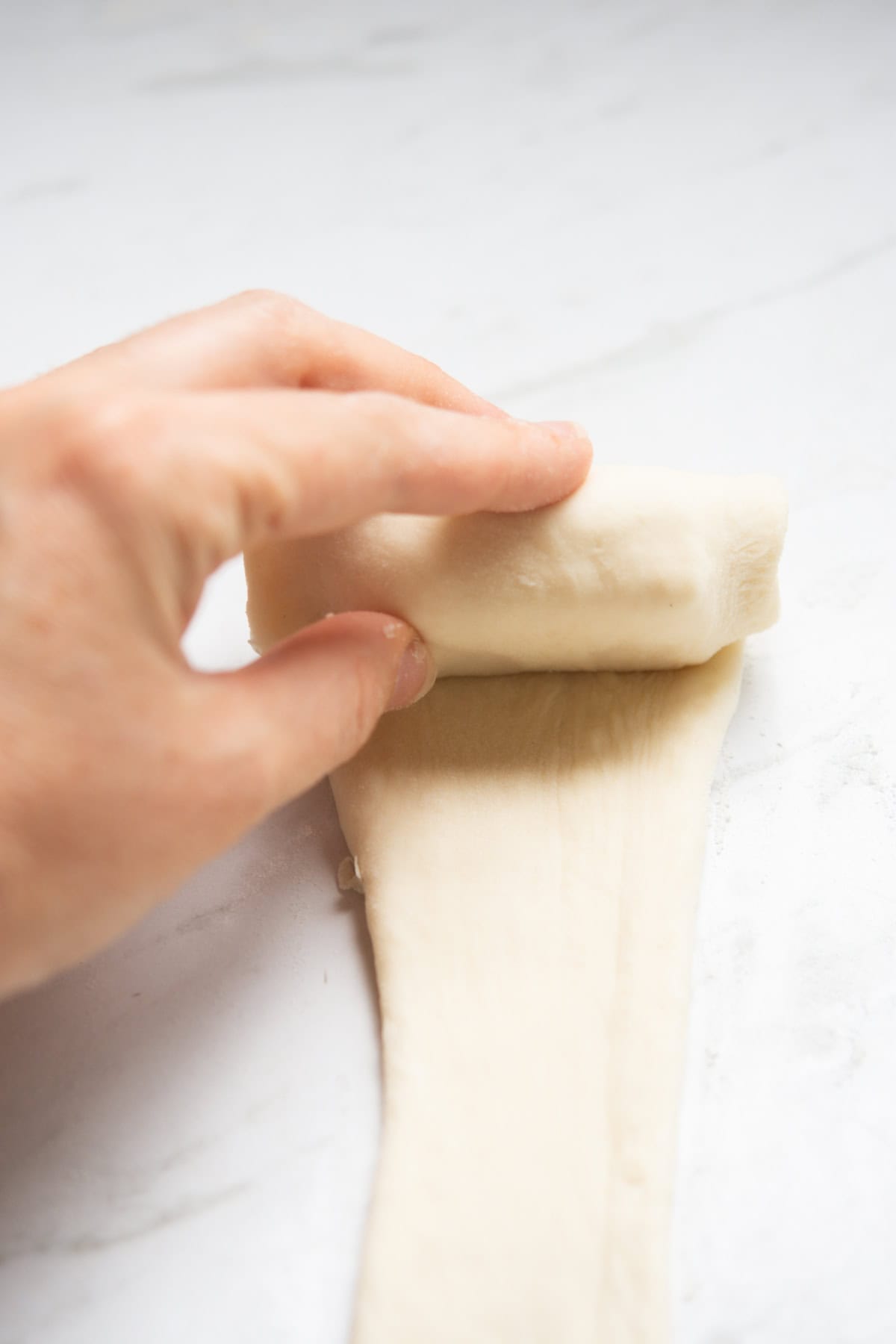 A hand rolls a piece of dough into a log shape on a white, lightly floured surface.