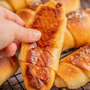A hand holding a golden-brown pastry with a caramelized top, with more similar pastries topped with coarse salt cooling on a wire rack in the background.