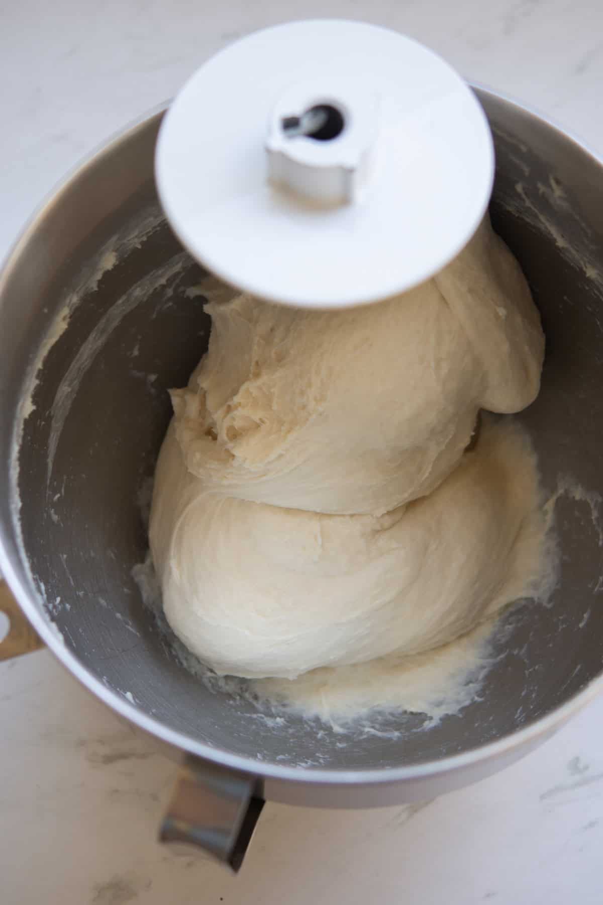 A stand mixer bowl filled with smooth, kneaded bread dough, attached to a dough hook on a white countertop.