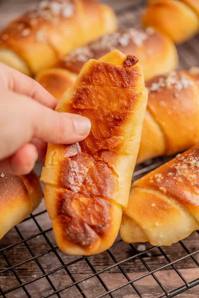 A hand holding a golden-brown pastry with a caramelized top, with more similar pastries topped with coarse salt cooling on a wire rack in the background.