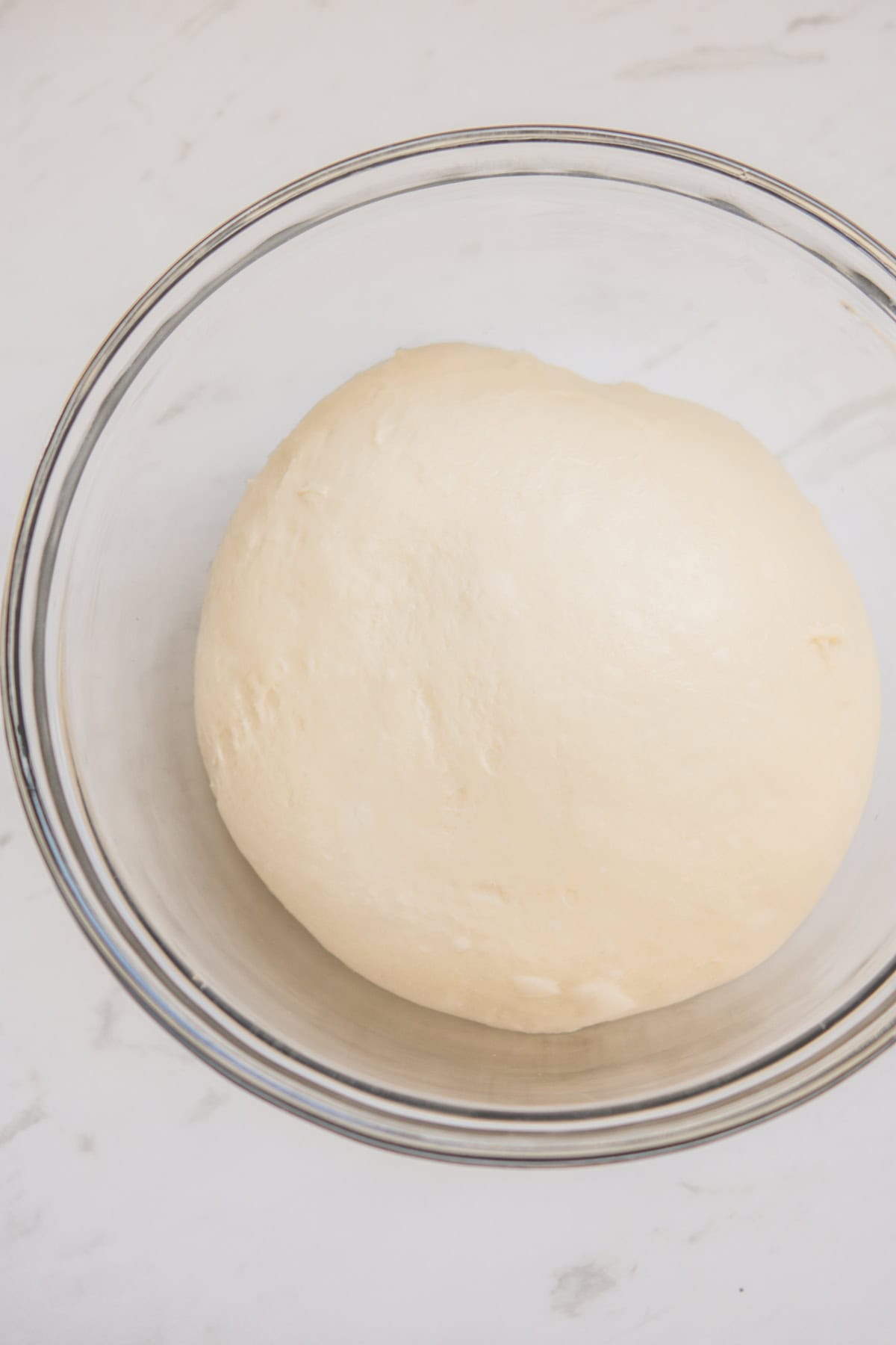 A smooth, round ball of dough resting in a clear glass bowl on a white marble surface.