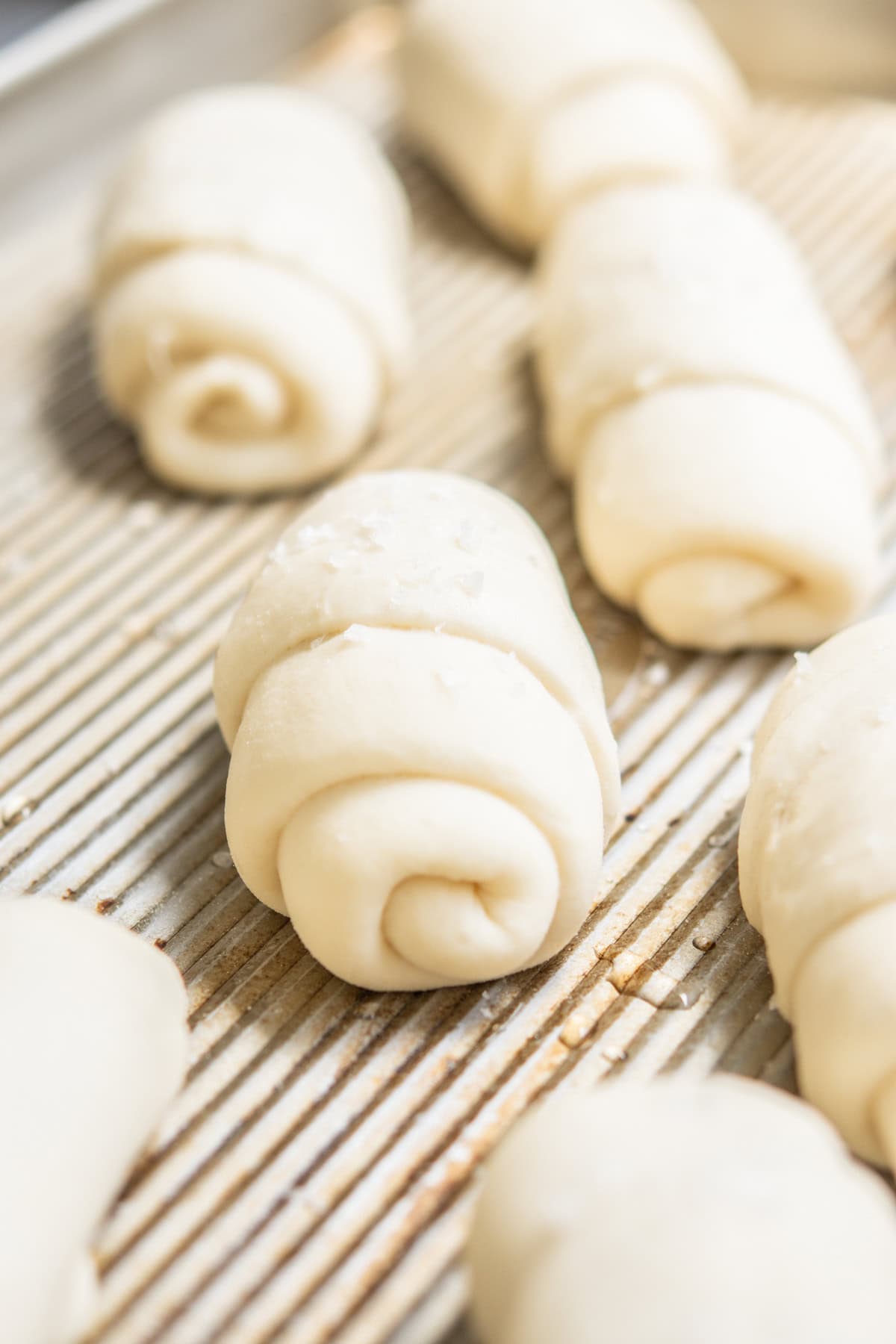 Close-up of several unbaked crescent rolls made from dough, arranged on a ridged baking sheet. The rolls are lightly dusted with flour and ready to be baked.