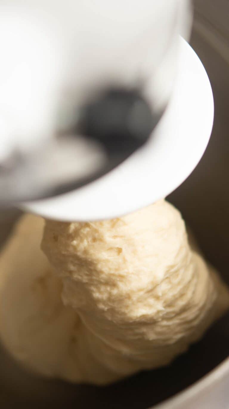 Close-up of bread dough being kneaded by a stand mixer with a dough hook attachment. The dough appears soft and elastic inside the metal mixing bowl.