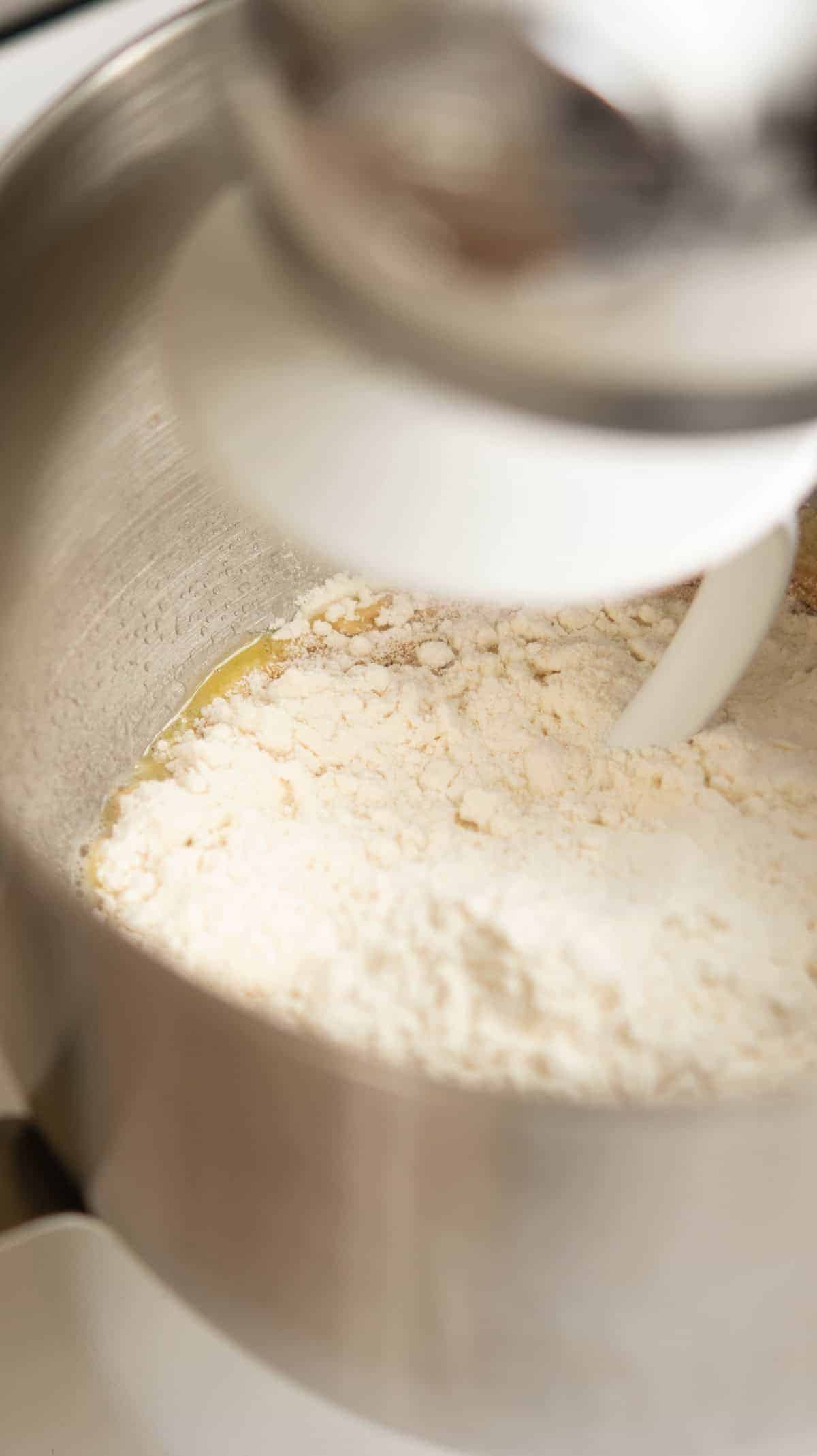 A close-up of flour and wet ingredients being mixed in a stand mixer bowl with a white dough hook attachment.