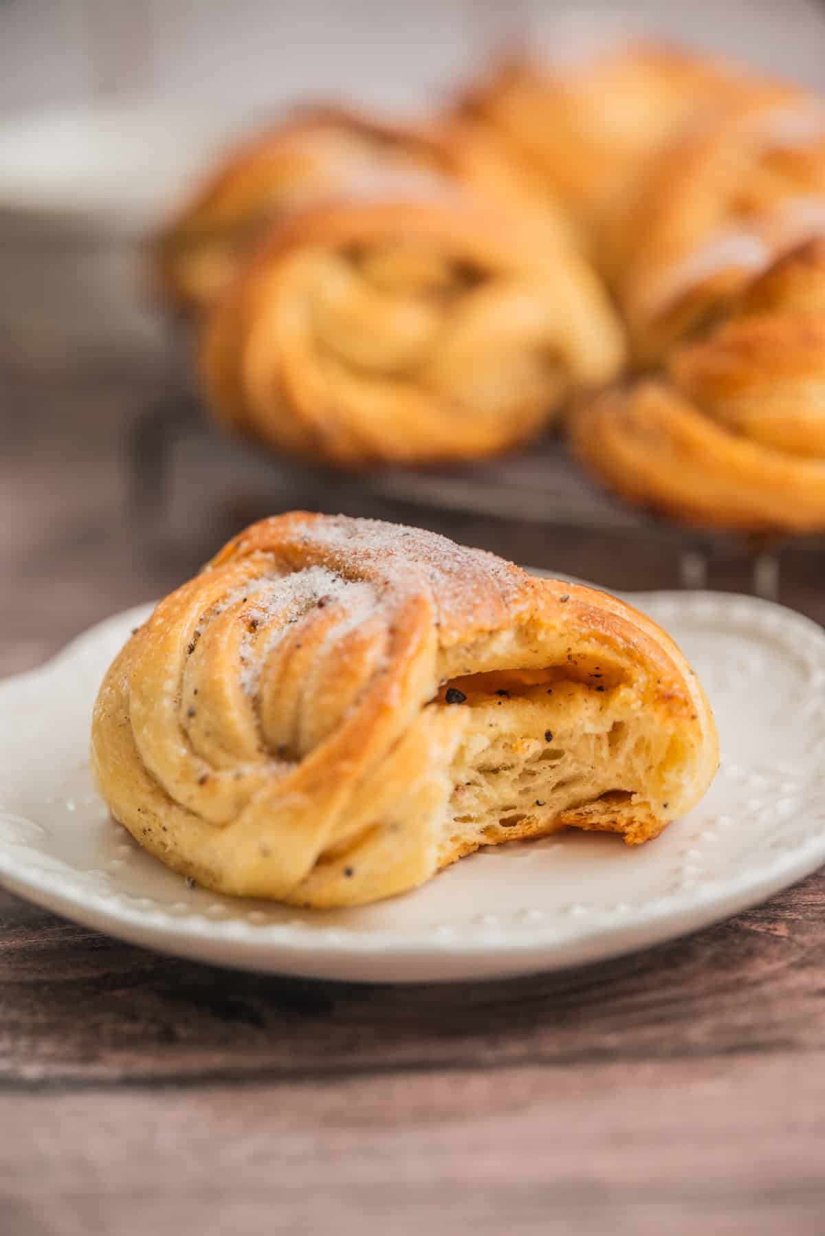 A close-up of a golden-brown sourdough cardamom bun lightly dusted with sugar, sits on a white plate. More pastries are blurred in the background.