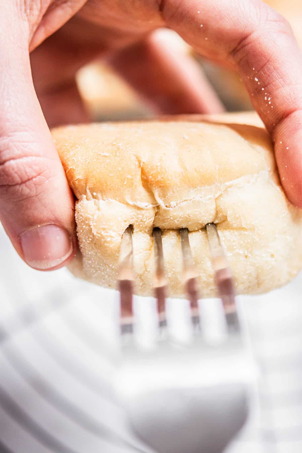 A close-up of a hand holding a soft bread roll while pressing the tines of a fork into its side, creating indentations on the bread’s surface.